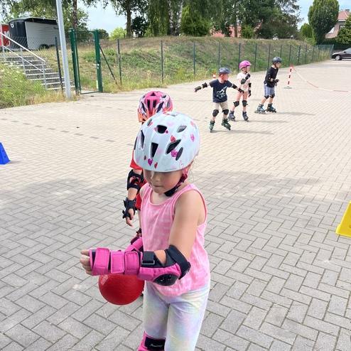 Ein M&auml;dchen mit pinkem Helm und Schutzausr&uuml;stung h&auml;lt einen roten Ball, w&auml;hrend andere Kinder in der Ferne skaten.