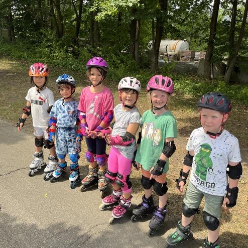 Sechs Kinder in Schutzhelmen und Schutzausr&uuml;stung stehen inline skaten auf einem gepflasterten Weg.