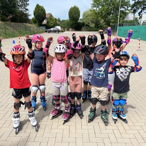Gruppe von zehn Kindern in Schutzausr&uuml;stung mit Rollschuhen, die sich freudig auf einem Platz versammeln.