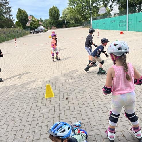 Gruppe von Kindern in Schutzkleidung beim Rollschuhlaufen auf einem asphaltierten Platz mit Verkehrsleitkegeln.