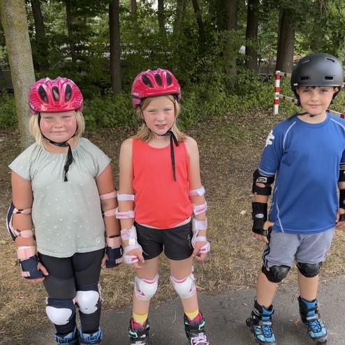 Drei Kinder in Schutzausr&uuml;stung und Helmen stehen auf einer Strecke zum Rollschuhlaufen im Freien.