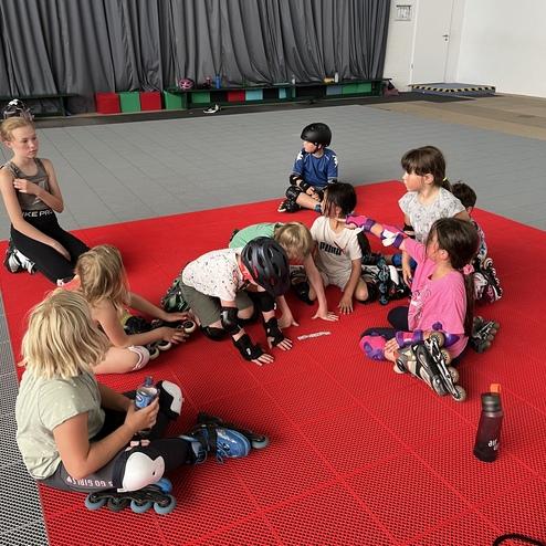 Gruppe von Kindern mit Inlinern sitzt auf einer roten Matte, w&auml;hrend sie sich um eine Ausr&uuml;stungssitzung versammeln.