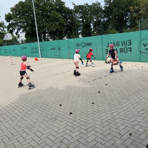 Kinder in Schutzkleidung &uuml;ben das Rollschuhlaufen auf einem gepflasterten Platz mit gr&uuml;ner Umz&auml;unung.