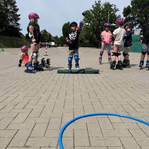 Kinder in Schutzausr&uuml;stung stehen auf einem Asphaltplatz, w&auml;hrend ein Junge auf einem Kissen mit einem Skateboard posiert.