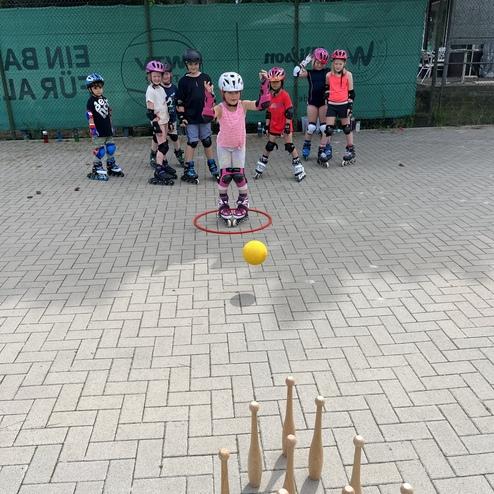 Kinder in Schutzausr&uuml;stung stehen hinter einer roten Markierung, w&auml;hrend ein Ball auf die Kegel vor ihnen gerollt wird.
