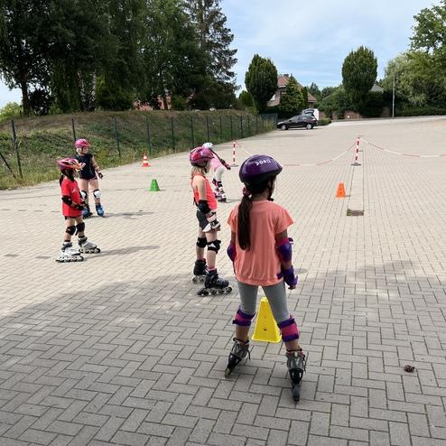Vier Kinder in Schutzausr&uuml;stung &uuml;ben auf Rollschuhen in einem abgegrenzten Bereich mit Kegeln und einer asphaltierten Fl&auml;che.