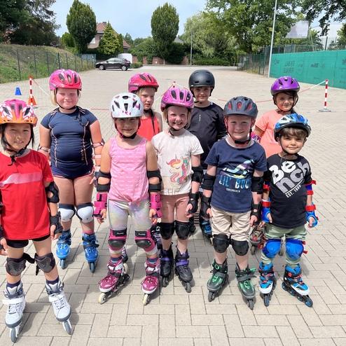 Gruppe von Kindern in Skate-Ausr&uuml;stung auf Inlineskates, l&auml;chelnd vor einer Asphaltfl&auml;che mit Verkehrsh&uuml;tchen.