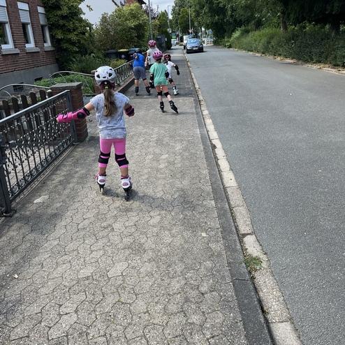 Vier Kinder in Schutzausr&uuml;stung fahren auf Rollerskates auf einem Gehweg entlang einer ruhigen Stra&szlig;e.