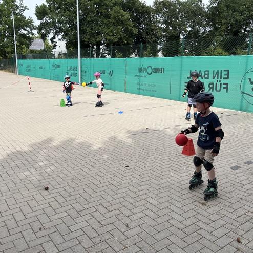Vier Kinder in Schutzkleidung &uuml;ben auf Rollskates auf einem Asphaltplatz mit Kegeln und einer roten Ball.