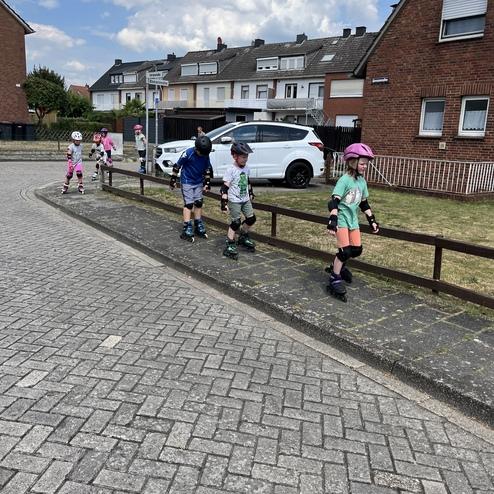 F&uuml;nf Kinder skaten auf einem gepflasterten Weg neben einem Zaun vor Wohnh&auml;usern, einige tragen Helme und Schutzausr&uuml;stung.