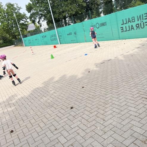 Zwei Kinder lernen in Schutzkleidung das Rollschuhlaufen auf einem gepflasterten Platz mit Markierungen.