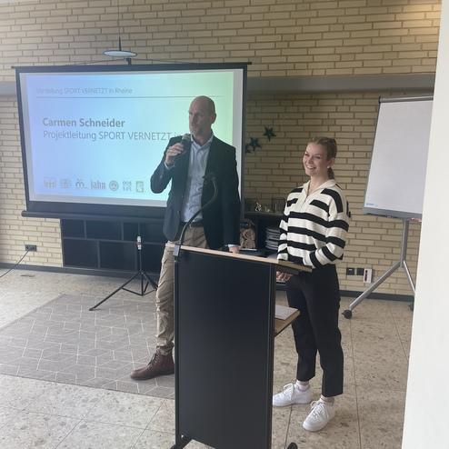 Mann mit Mikrofon spricht vor Publikum, während eine Frau mit Streifenpulli an einem Podium steht. Bildschirm im Hintergrund.