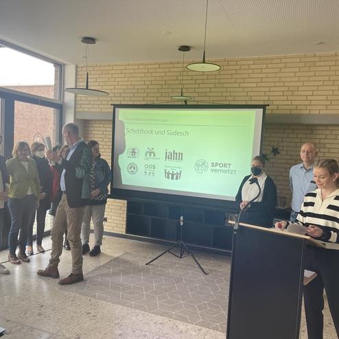 Gruppenszene in einem Seminarraum: Personen stehen vor einer Leinwand mit Logos, während eine Frau spricht.