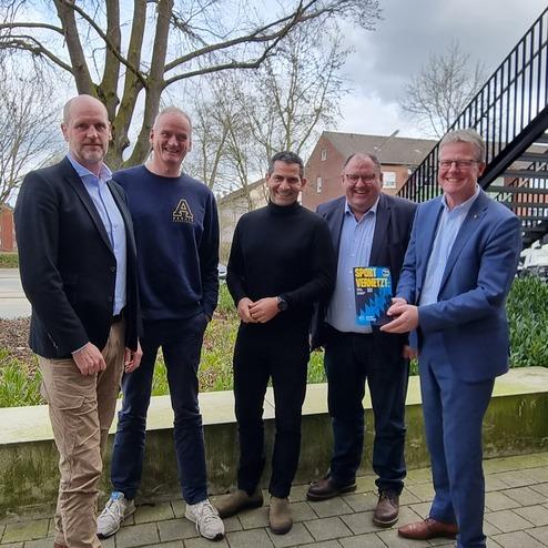 Fünf Männer stehen auf einem Gehweg vor einem Gebäude, einer hält ein Buch in der Hand, Bäume im Hintergrund.