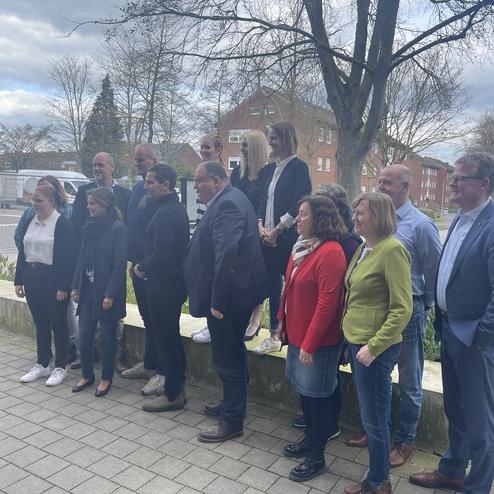 Gruppe von 12 Personen in formeller Kleidung steht auf einem Gehweg vor einem Gebäude mit Bäumen im Hintergrund.