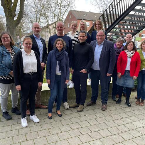 Gruppenfoto von 18 Personen, die auf einem Gehweg vor einem Gebäude stehen, umgeben von Bäumen und einem Fahrrad.