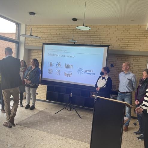 Gruppenszene mit Personen, die vor einer Präsentation zu „Schulbuch und Sudiesch“ in einem modernen Raum stehen.