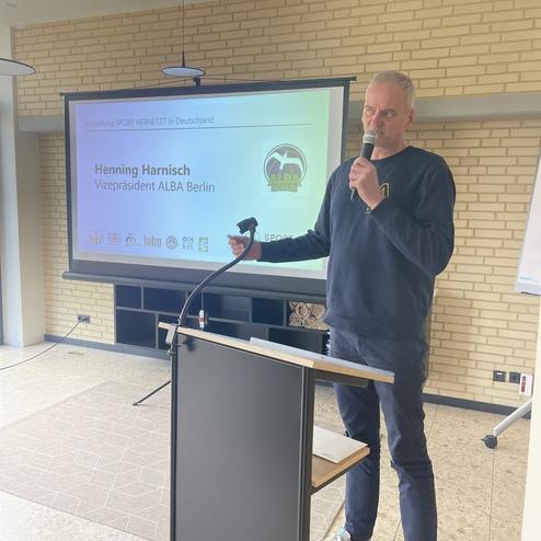 Mann spricht am Rednerpult während einer Präsentation mit Bildschirm im Hintergrund zu "Sport vernetzt in Deutschland".