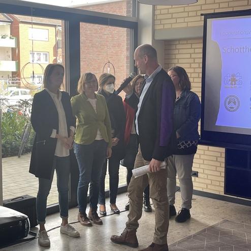 Gruppenszene mit fünf Personen in einem Raum; ein Mann spricht ins Mikrofon, während die anderen ihm zuhören.