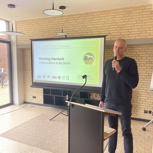 Mann spricht am Rednerpult vor einer Präsentation über die Zusammenarbeit im Sport, im Hintergrund ein Fenster und Fahrräder.
