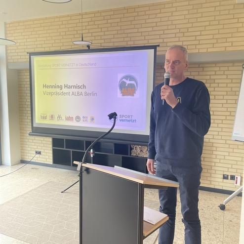 Mann spricht am Rednerpult vor einer Präsentation mit dem Titel „SPORT vernetzt“ im Hintergrund.