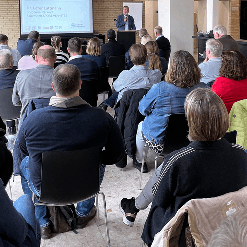 Dr. Peter Lüttmann spricht vor einem Publikum in einem Konferenzraum mit Stühlen und Projektorleinwand.