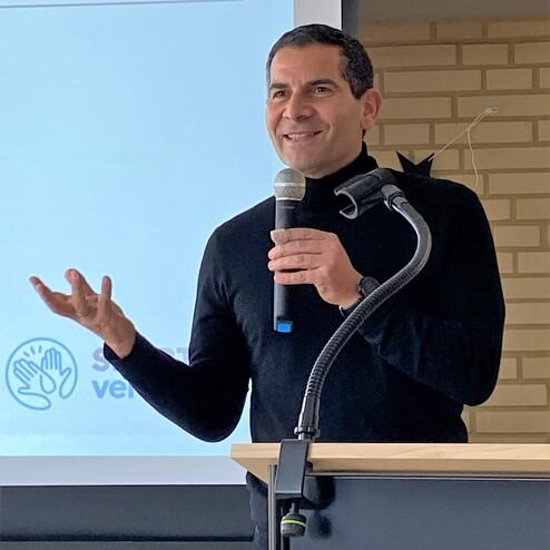 Mann in schwarzem Rollkragenpullover spricht lächelnd ins Mikrofon vor einem Bildschirm und Podium.