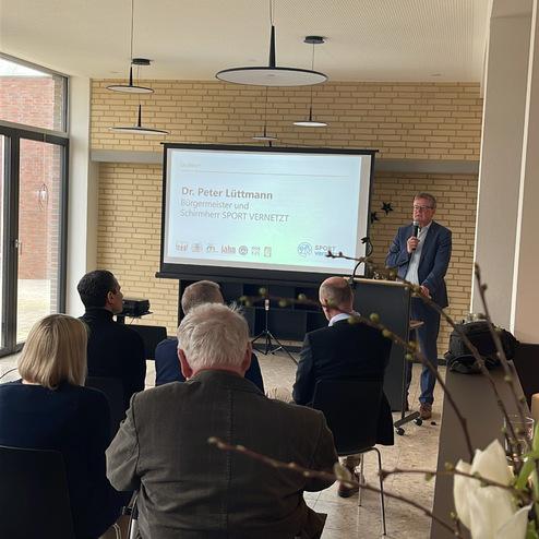 Dr. Peter Lüttmann spricht vor Publikum in einem modernen Raum, im Hintergrund eine Präsentation über "SPORT VERNETZT".