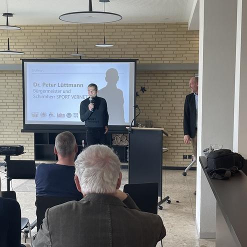 Dr. Peter Lüttmann spricht bei einer Veranstaltung, während ein Bildschirm im Hintergrund Informationen zeigt.