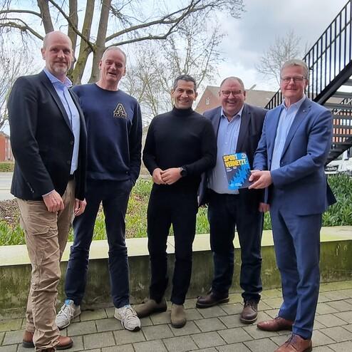 Fünf Männer stehen in einem Gartenbereich, sie lächeln und halten gemeinsam ein Buch in der Hand.