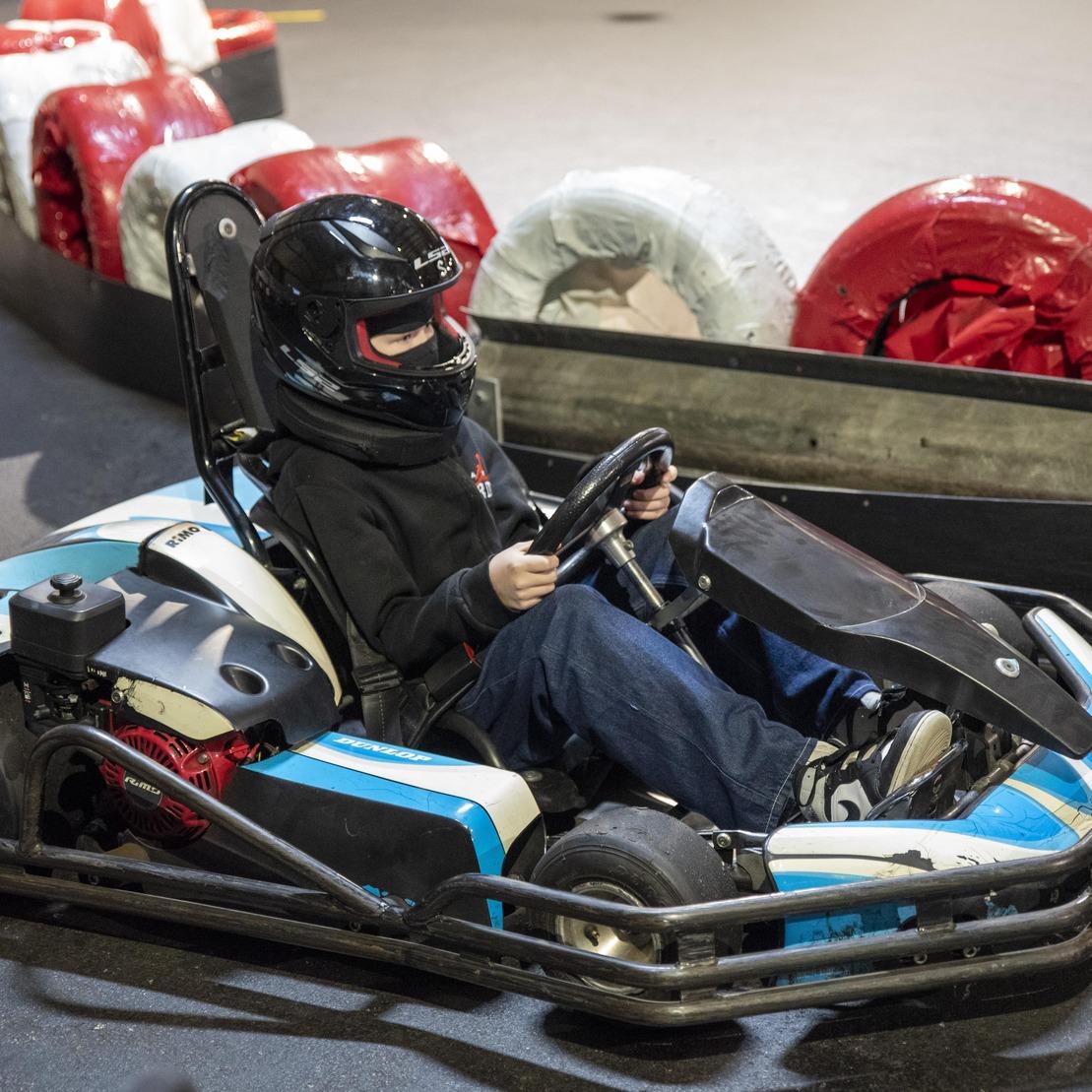 Ein Kind sitzt im Kartsport-Anzug in einem Go-Kart auf einer Rennbahn und h&auml;lt das Lenkrad.