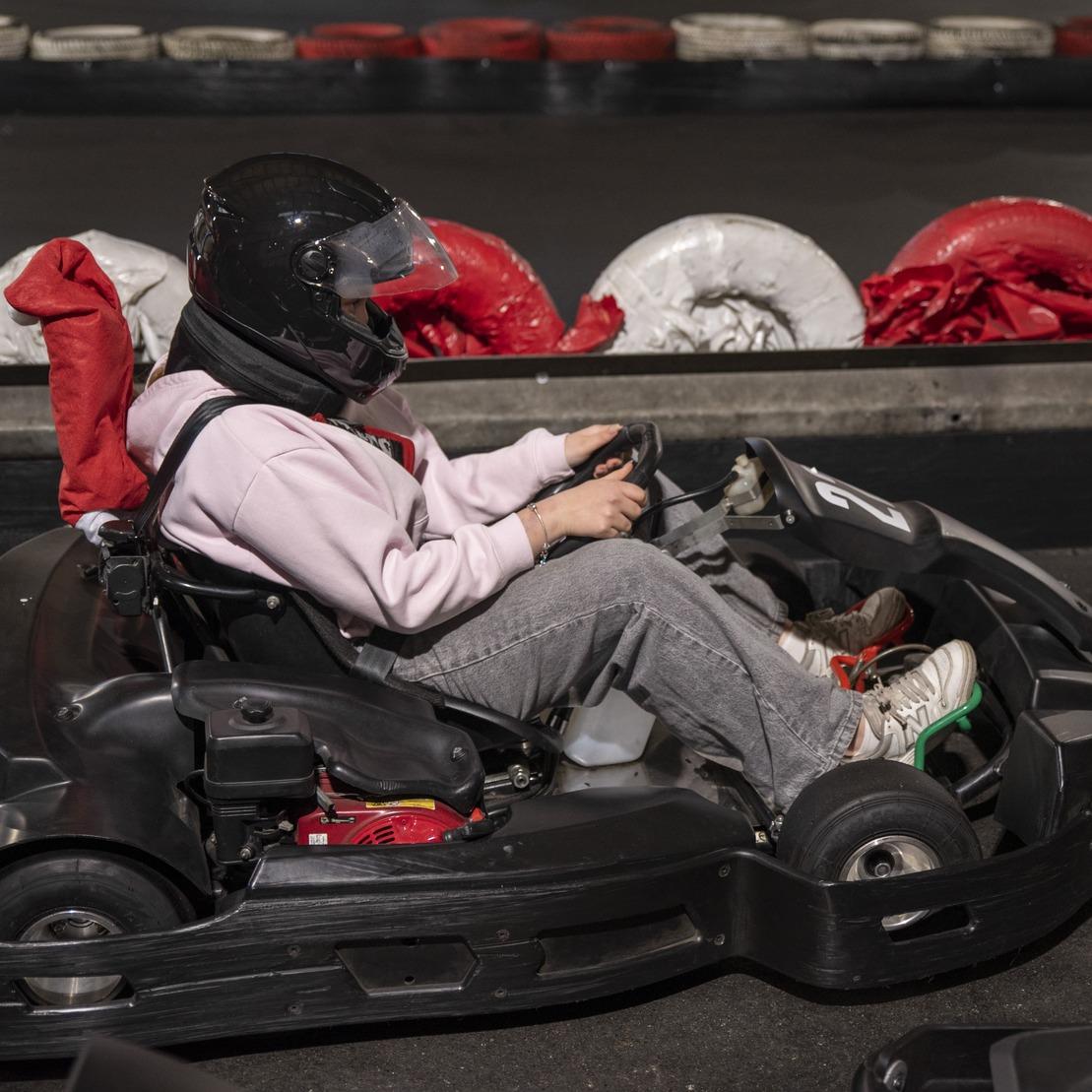 Person in einem schwarzen Kart, tr&auml;gt Helm und Schutzkleidung, sitzt auf einer Karts-Bahn mit rot-wei&szlig;en Reifen im Hintergrund.