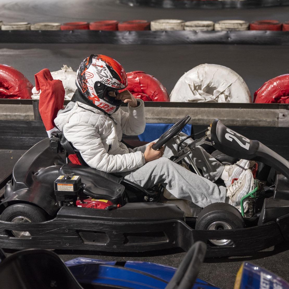 Junger Fahrer in einem Go-Kart, sitzend, mit Helm und wei&szlig;er Jacke, auf einer Rennstrecke mit rot-wei&szlig;en Reifen.