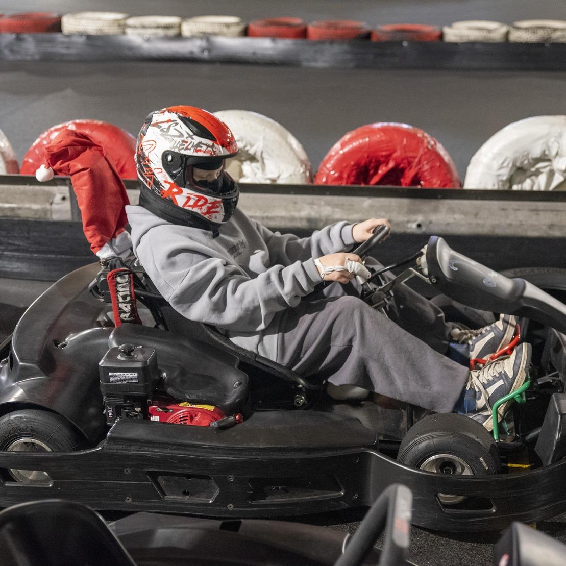 Ein Kind in einem Rennanzug sitzt in einem Kart auf einer Kartbahn, umgeben von Reifen und einem schwarzen Boden.