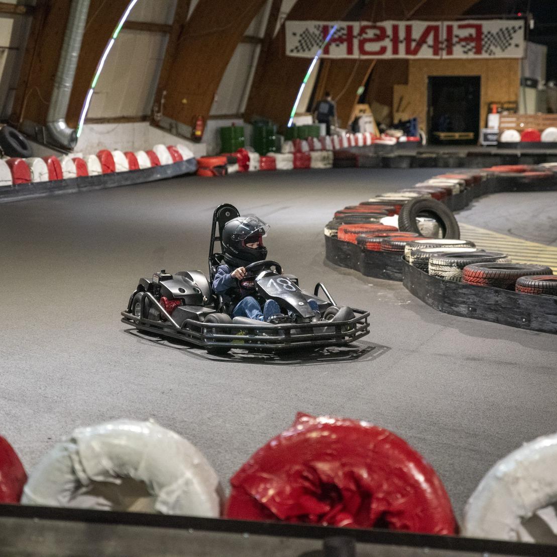 Ein Kartfahrer in einem schwarzen Go-Kart kurvt auf einer Rennstrecke mit roten und wei&szlig;en Reifenstapeln.