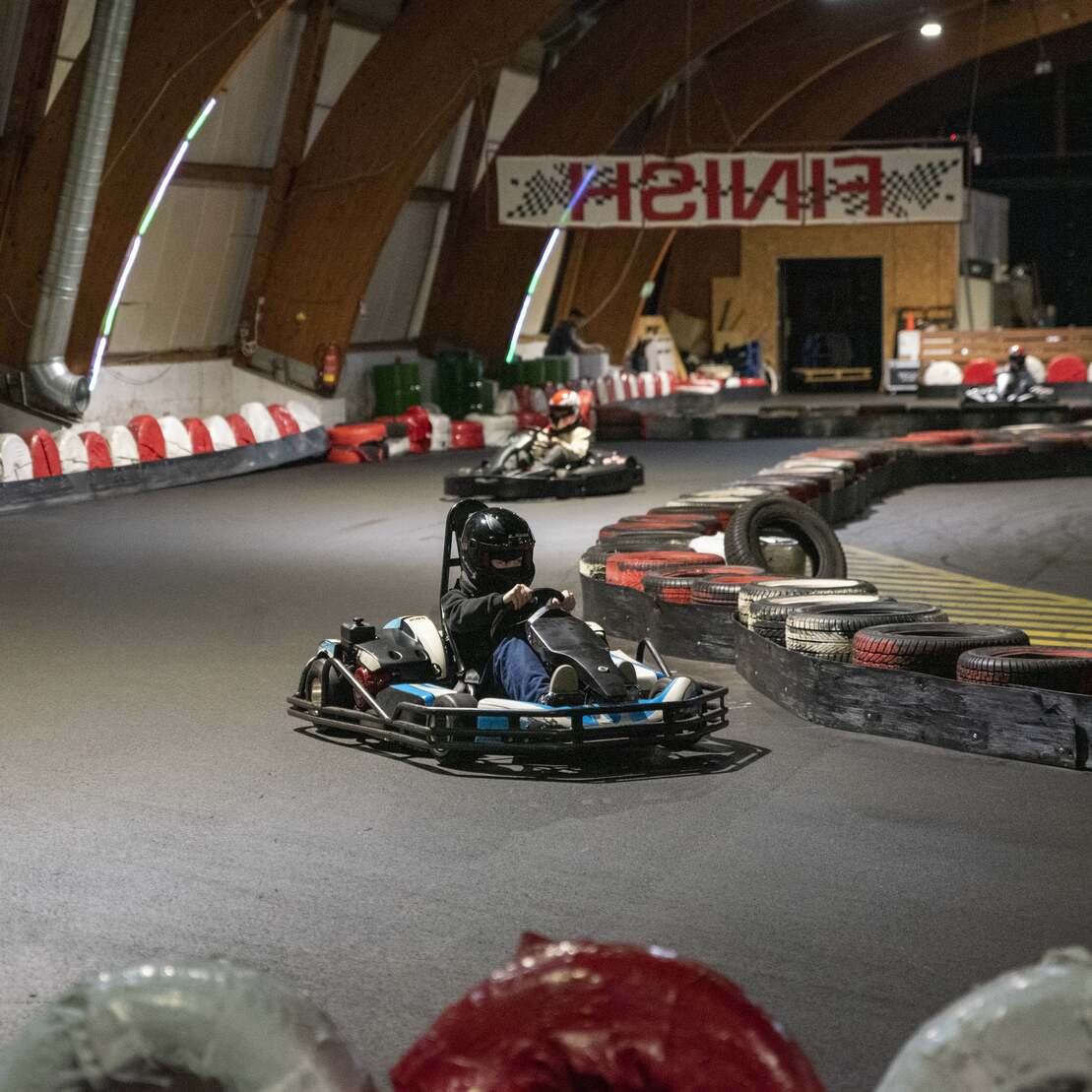 Go-Kart auf einer Innenbahn mit kurvenreicher Strecke und Zielbanner im Hintergrund. Fahrer mit Helm beim Rasen.