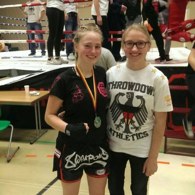 Zwei Frauen stehen l&auml;chelnd vor einem Boxring, die Sportlerin tr&auml;gt eine Medaille und Kampfbekleidung.