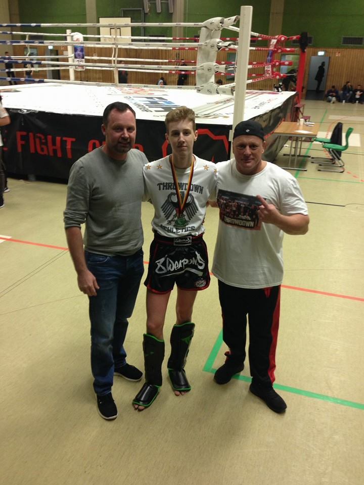 Drei Personen posieren in einem Sportumfeld. Der junge K&auml;mpfer tr&auml;gt eine Medaille und ist in Kampfausr&uuml;stung.