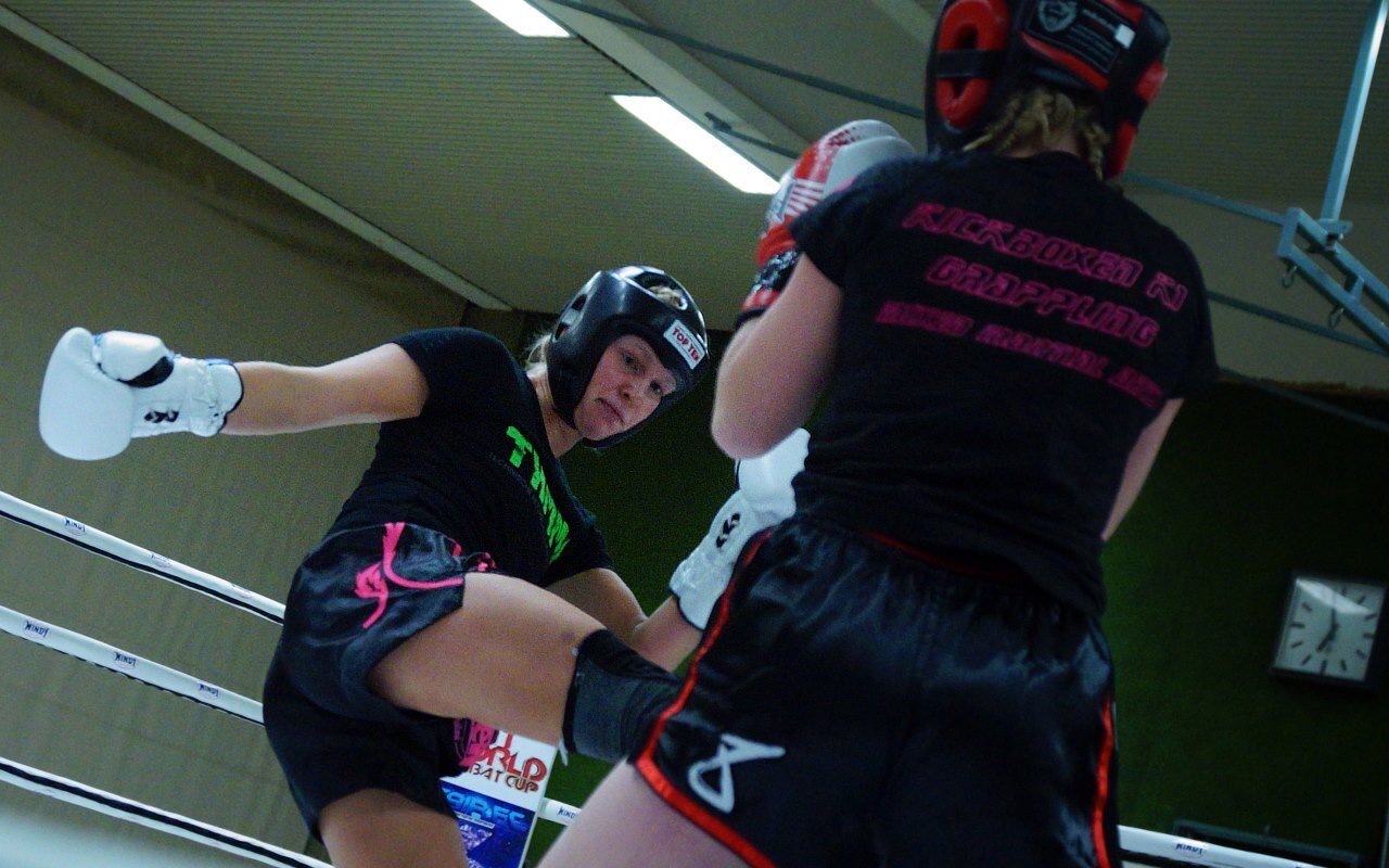 Kampfsportlerinnen im Ring, eine mit Kicktechnik, die andere im Defensive, beide in Schutzausr&uuml;stung.