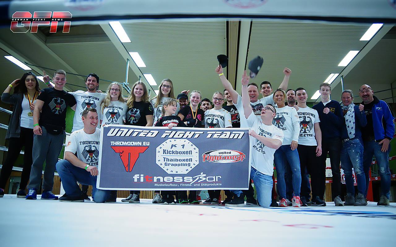Gruppe von K&auml;mpfern und Unterst&uuml;tzern posiert gemeinsam vor einem Banner des United Fight Team in einer Sporthalle.