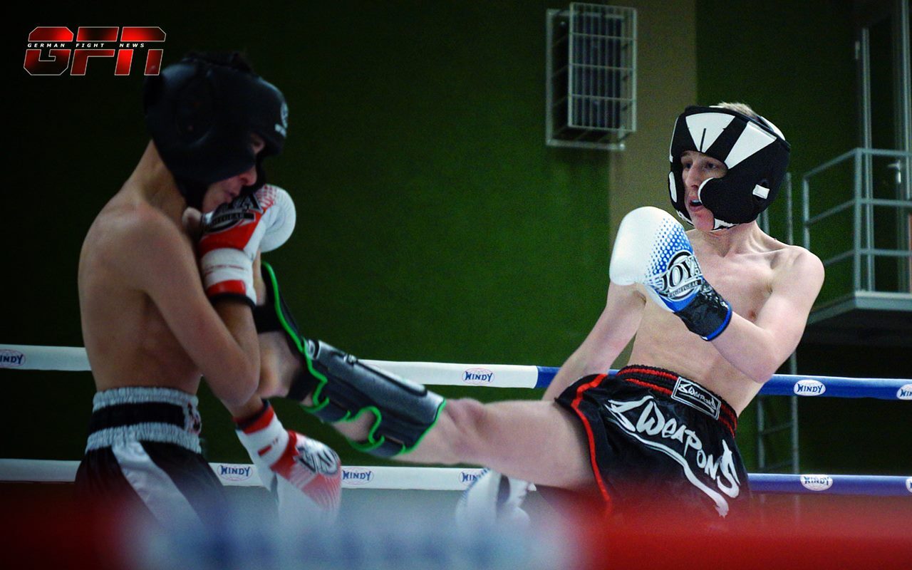 Zwei Boxer im Ring; einer trifft den Gegen&uuml;ber mit einem hohen Kick, beide tragen Schutzausr&uuml;stung.