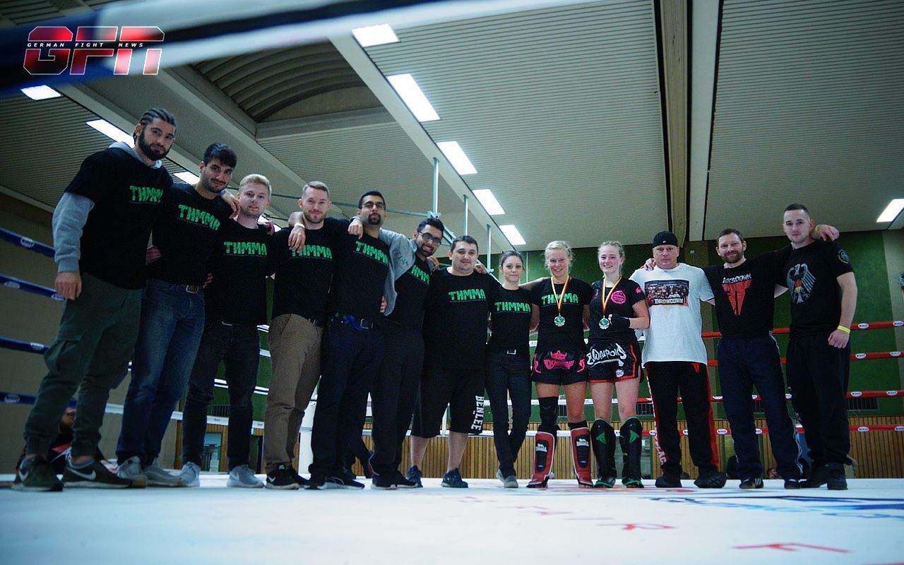 Gruppe von zehn Personen in Kampfsport-T-Shirts auf einer Boxringfl&auml;che, l&auml;chelnd und posierend nach einem Wettkampf.