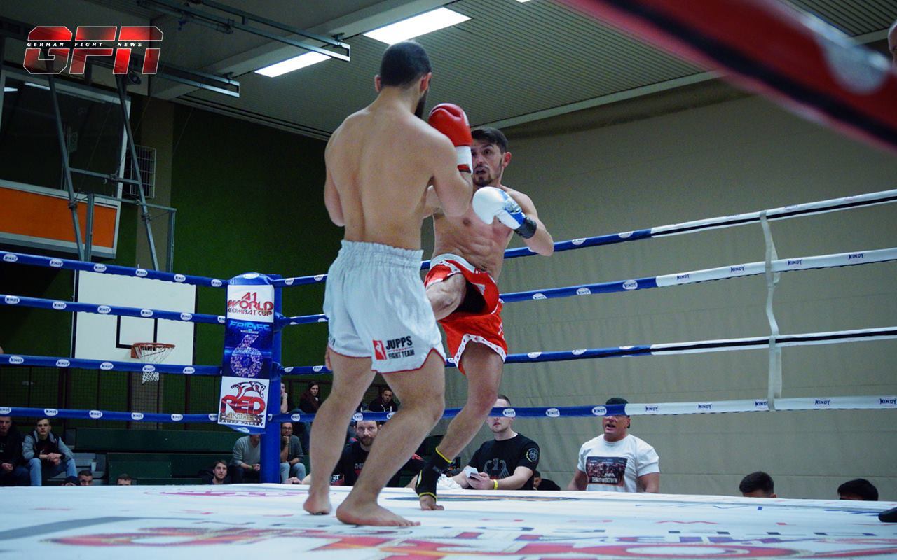 Zwei K&auml;mpfer im Ring, einer f&uuml;hrt einen Kick aus, w&auml;hrend der andere ausweicht. Zuschauer im Hintergrund sind sichtbar.