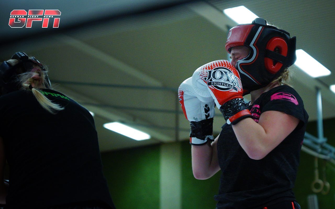 Zwei Frauen im Boxtraining, eine tr&auml;gt Schutzausr&uuml;stung und Boxhandschuhe, die andere steht im Hintergrund.