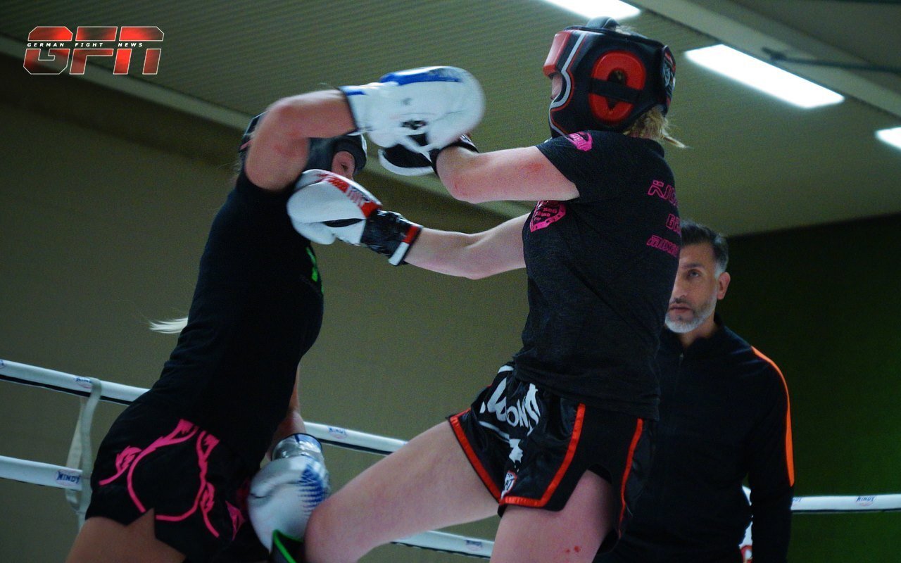 Zwei Frauen im Boxring w&auml;hrend eines Wettkampfs; eine Frau trifft die andere mit einem Schlag, ein Trainer beobachtet.