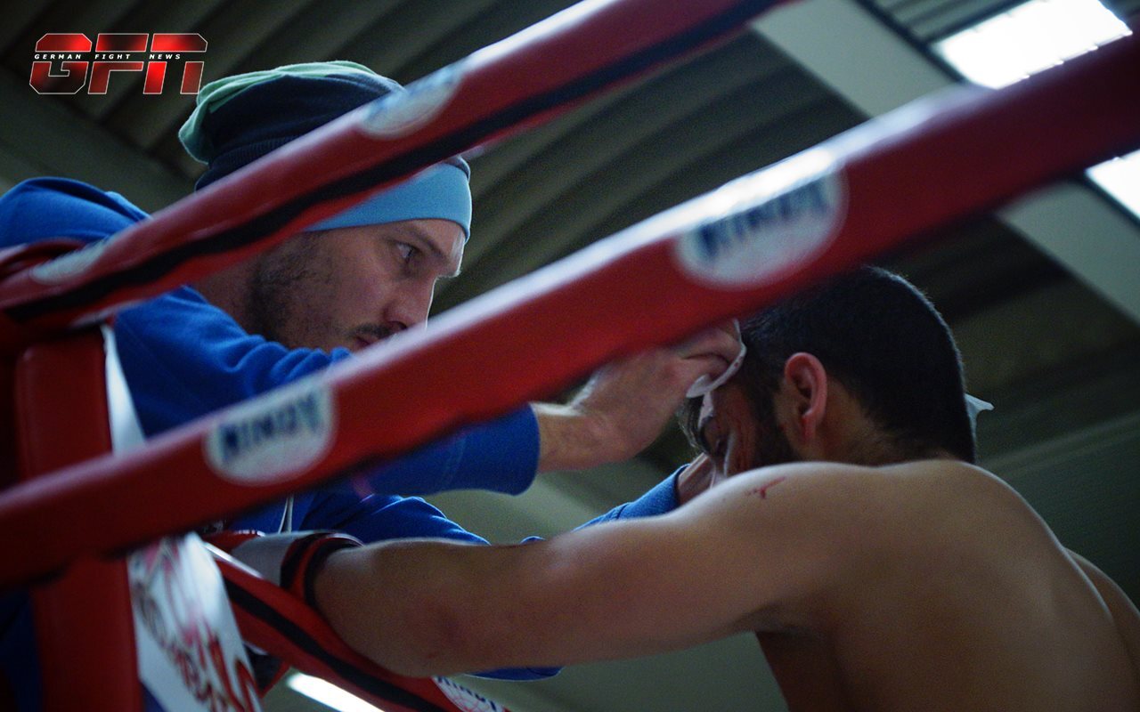 Trainer reinigt das Gesicht eines K&auml;mpfers w&auml;hrend einer Pause im Boxring, umgeben von roten Ringen.