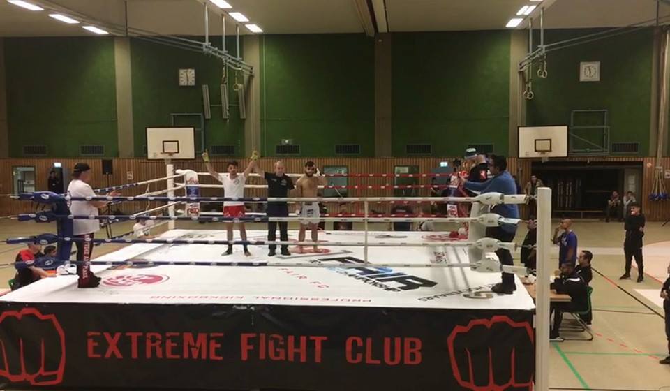 Zwei K&auml;mpfer stehen im Ring des Extreme Fight Clubs, w&auml;hrend Zuschauer und Offizielle zuschauen.