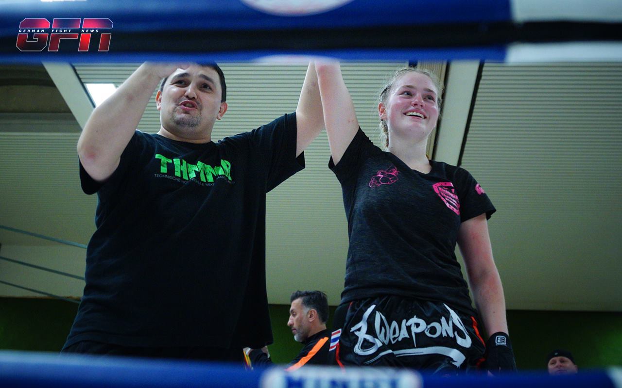 Trainer hebt die Hand einer siegreichen K&auml;mpferin nach einem Boxkampf in einer Sporthalle.