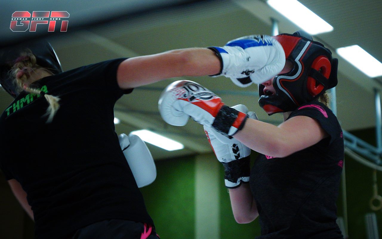 Zwei Sportlerinnen im Boxtraining, eine schl&auml;gt mit einem Handschuh auf die andere, beide tragen Schutzhelme.