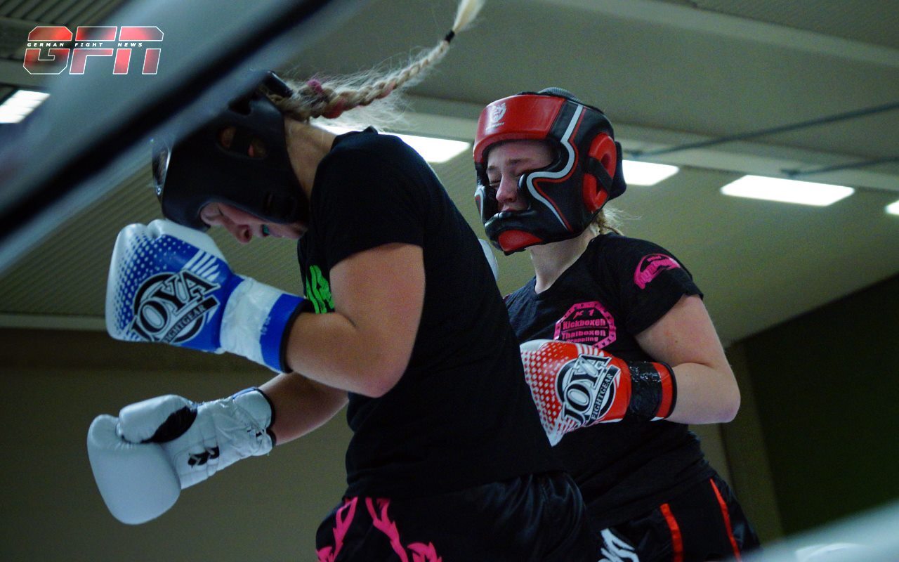 Zwei Boxerinnen im Sparring mit Schutzausr&uuml;stung in einer Sporthalle, eine schl&auml;gt mit einem Handschuh zu.